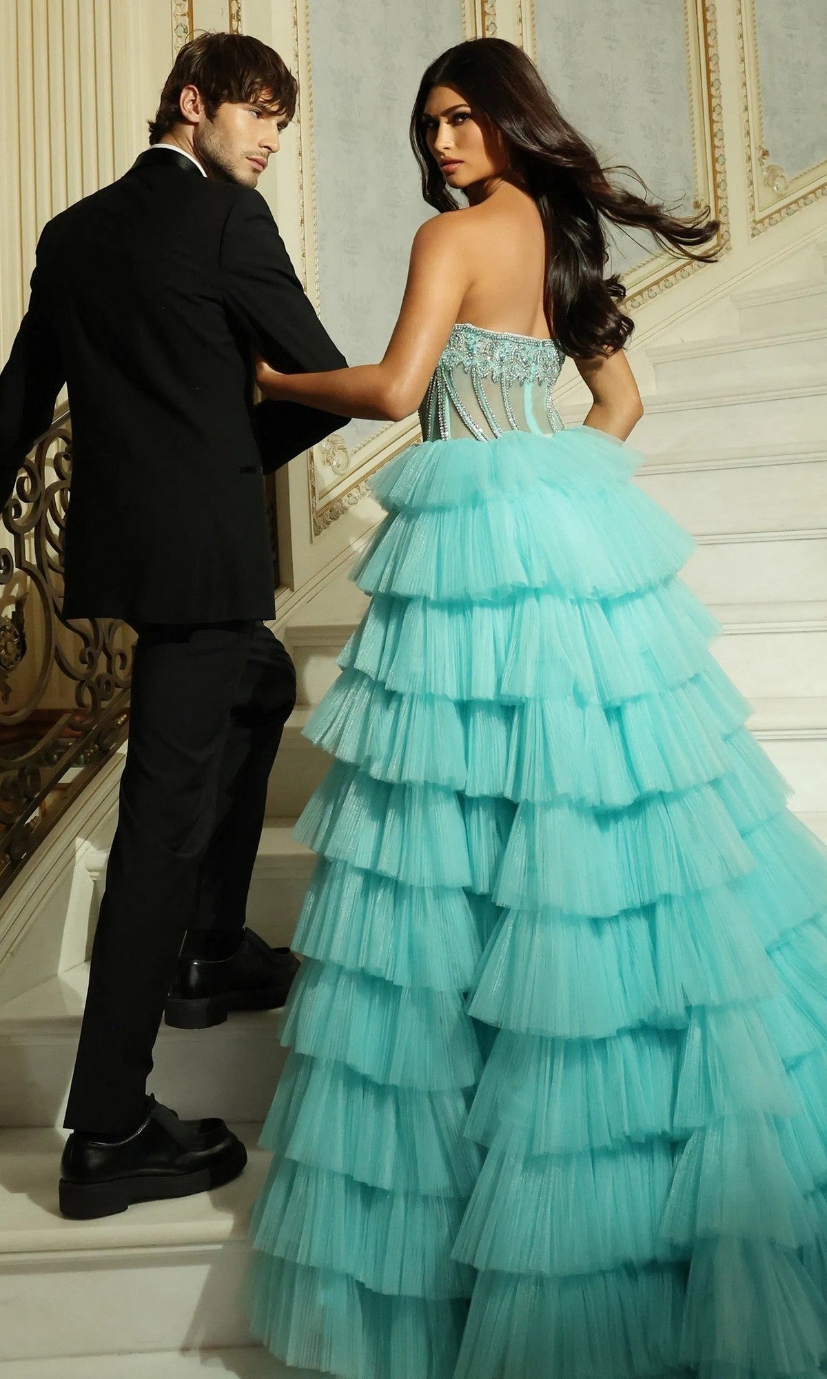 A woman in the Jovani 44134 long prom dress and a man in a black suit walk up a grand staircase, glancing back over their shoulders.
